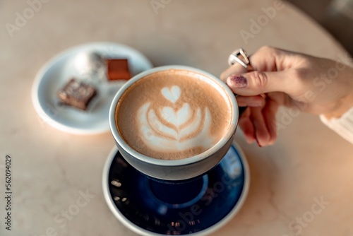 A cup of fresh cappuccino coffee in the hands of a woman on a fashionable background of a white marble table, next to a plate with sweets. Coffee addiction. Top view, flat lay.
