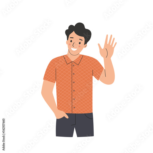Young man waives hand in hello gesture while smiling cheerfully and holding hand in pocket