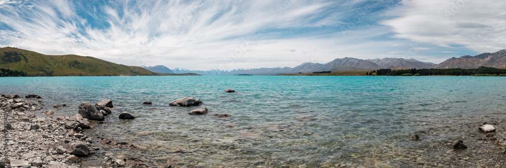 Panoramic view of wispy clouds and blue skies over the azure blue water of Lake Tekapo in New Zealand with snow capped mountains in the distance and rocky shore in the foreground