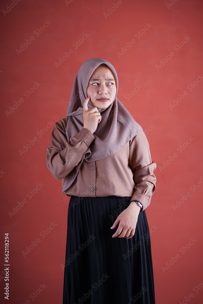 Beautiful Asian woman in brown shirt and hijab showing thinking expression with hand on cheek on brown background Beautiful Asian woman in brown shirt and hijab showing thinking expression with hand on cheek on brown background
