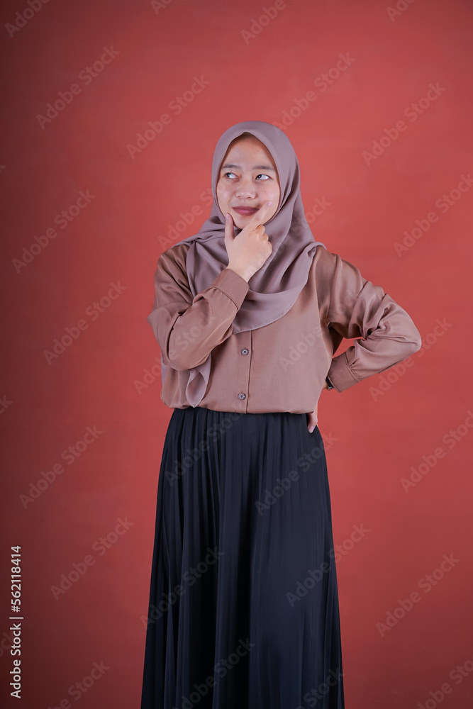 Beautiful Asian woman in brown shirt and hijab showing thinking expression with hand on chin on brown background Beautiful Asian woman in brown shirt and hijab showing thinking expression with hand on chin on brown background
