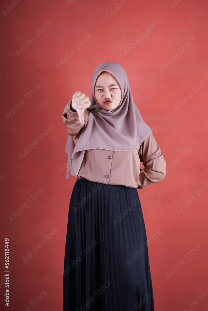 Beautiful Asian woman in brown shirt and hijab shows upset expression with thumbs down on brown background Beautiful Asian woman in brown shirt and hijab shows upset expression with thumbs down on brown background