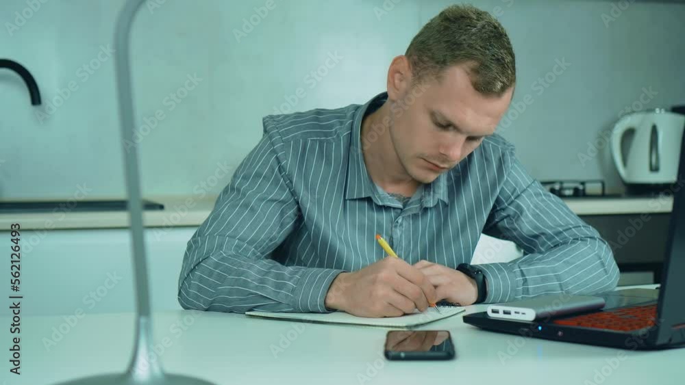 Focused business man entrepreneur writing on paper doing research. Young male professional using computer sitting at home office desk. Busy worker freelancer working at kitchen in financial sphere Focused business man entrepreneur writing on paper doing research. Young male professional using computer sitting at home office desk. Busy worker freelancer working at kitchen in financial sphere