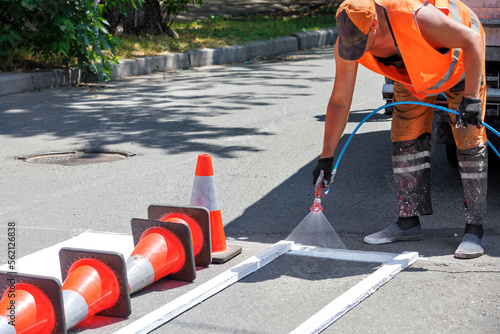 A road worker in orange overalls paints white road markings at a pedestrian crossing on a summer day.