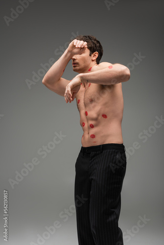 man in black pants with red lip prints on shirtless torso obscuring face while posing on grey background.