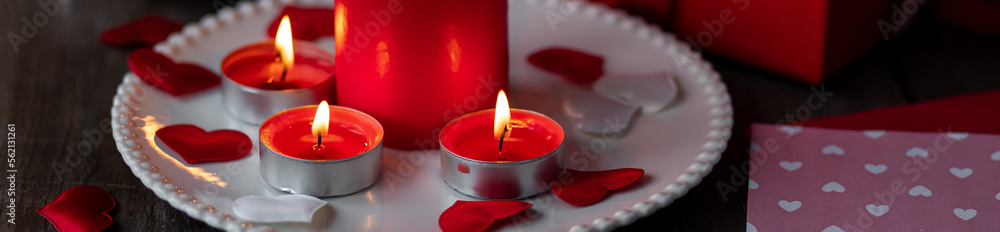 Saint Valentine's Day celebration. Red burning candles, hearts, gift box, postcard on dark wooden background. Happy holiday. Table decor for festive dinner, romantic atmosphere. Banner Saint Valentine's Day celebration. Red burning candles, hearts, gift box, postcard on dark wooden background. Happy holiday. Table decor for festive dinner, romantic atmosphere. Banner