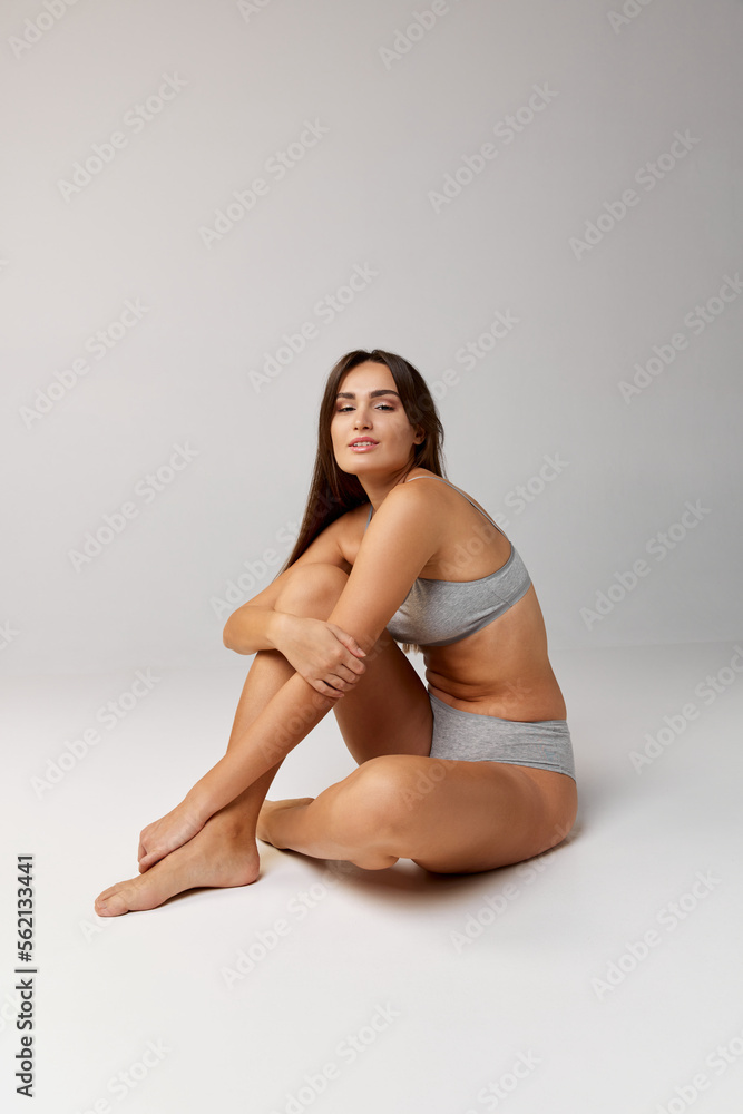 Portrait of young beautiful girl sitting on floor and posing in underwear over grey studio background. Self-care. Concept of body and skin care, fitness Portrait of young beautiful girl sitting on floor and posing in underwear over grey studio background. Self-care. Concept of body and skin care, fitness