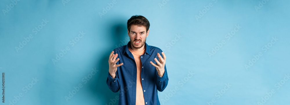 Annoyed and angry man raising hands and clench teeth outraged, stare pissed-off at camera, going to kill someone, standing on blue background - obrazy, fototapety, plakaty 