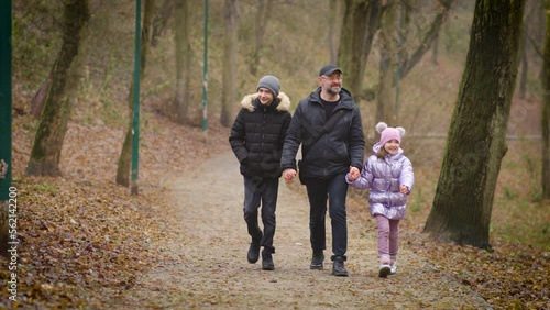 Front view of adult father and little fanny daughter with teenager son walking along autumn forest path cloudy day holding hands and talking. Concept of joint family pastime, parenting and upbringing