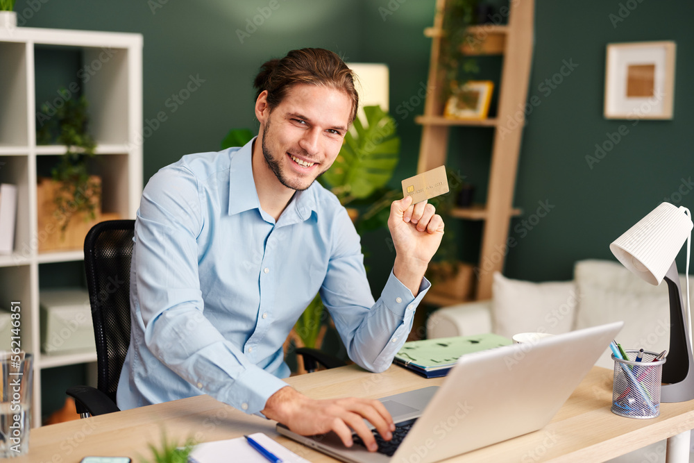 Portrait of young man sitting at a table with a laptop in front of him and holds a credit card in his hand