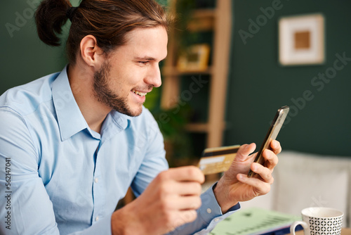 Portrait of young man sits at a table with a smartphone and credit card in his hand