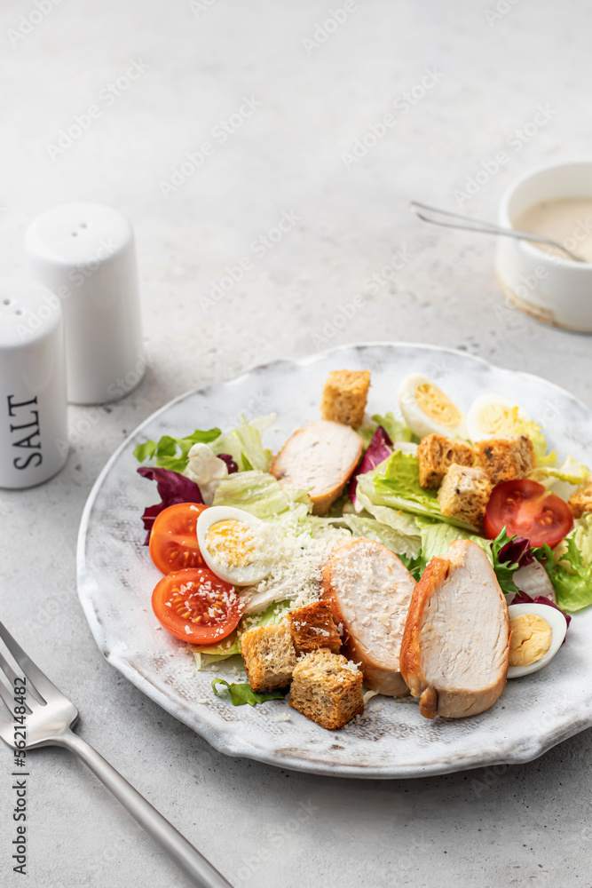 Caesar salad. Healthy salad made with chicken fillet, cherry tomatoes, iceberg lettuce, parmesan cheese and croutons on light grey background with cutlery. Light diet dinner, text space Caesar salad. Healthy salad made with chicken fillet, cherry tomatoes, iceberg lettuce, parmesan cheese and croutons on light grey background with cutlery. Light diet dinner, text space