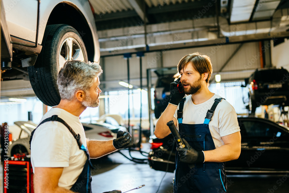 Modern workshop. Young beard male professional technician mechanic man wears denim blue overalls t-shirt use talk mobile cell phone stand near car lift. Modern workshop. Young beard male professional technician mechanic man wears denim blue overalls t-shirt use talk mobile cell phone stand near car lift.