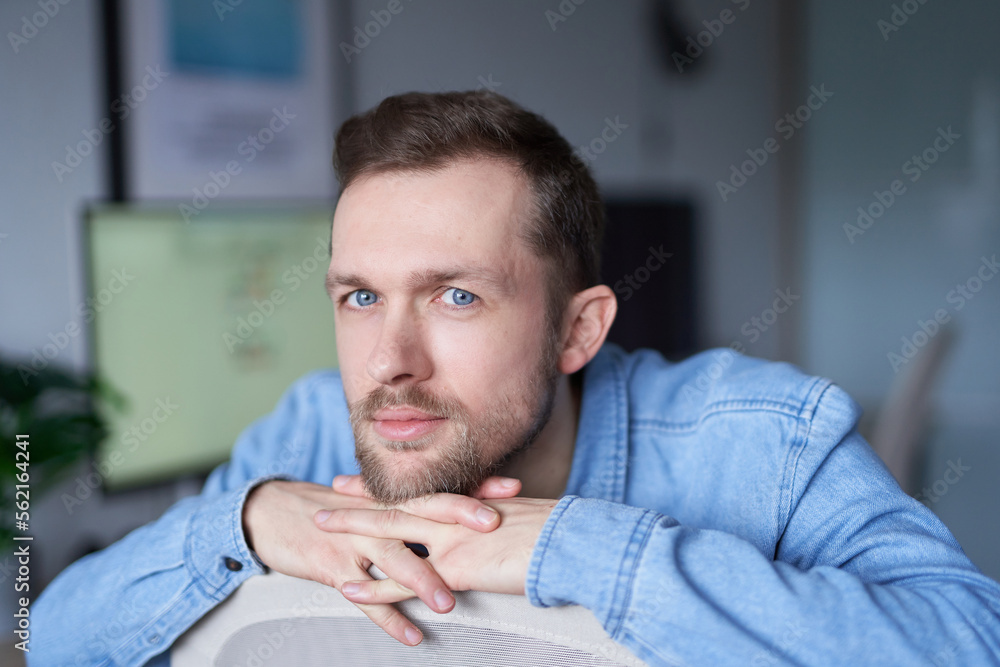 Young handsome man in denim shirt, positive emotion with computer monitor on backgroundSerious bearded male software engineer sitting at working place in office looking at camera. High quality image