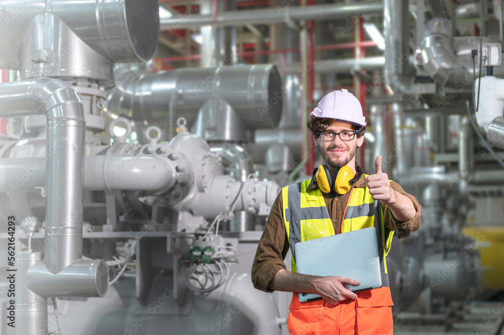 A male engineer in a portrait holding a laptop while a show thump up inside a factory. A male engineer in a portrait holding a laptop while a show thump up inside a factory.