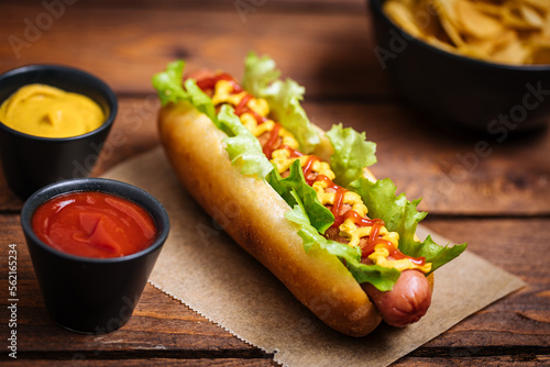 Hot dog with mustard, ketchup and green salad in bun, served with potato chips on a rustic wooden board. Fast food and delicious.