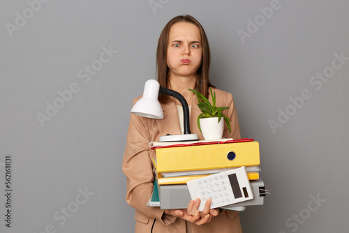 Portrait of dismissed crazy woman wearing stylish beige jacket holding office stuff posing isolated over gray background, expressing sadness and sorrow, lost job, keeps eyes crossed. photo