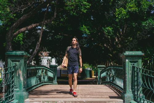 Young Latin man with long hair and wearing a smart watch passing a wooden bridge in a city park with a yoga mat on his back.