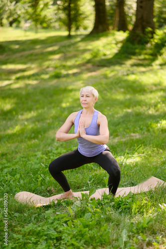 Middle-aged woman doing yoga in sportswear outdoors in a city park in Namaskarasana - Seated Prayer Pose. Concept of stretching, pilates, healthy lifestyle.