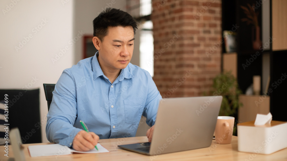 Confident middle aged asian man making notes, working on laptop in home office interior, panorama, free space Confident middle aged asian man making notes, working on laptop in home office interior, panorama, free space