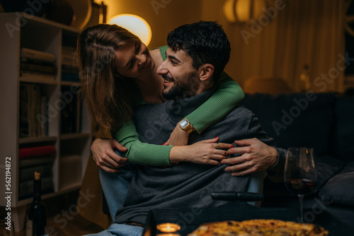 Smiling woman hugging her boyfriend on the couch from behind in the living room.