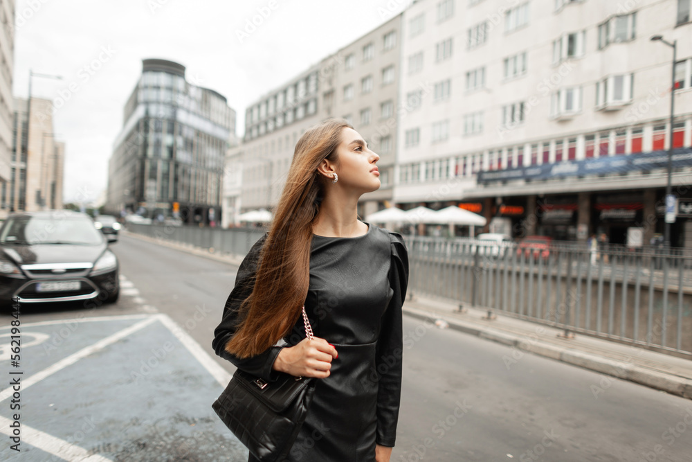 Beautiful business fashion woman in stylish black dress with fashionable black leather handbag walks near a road in the modern city