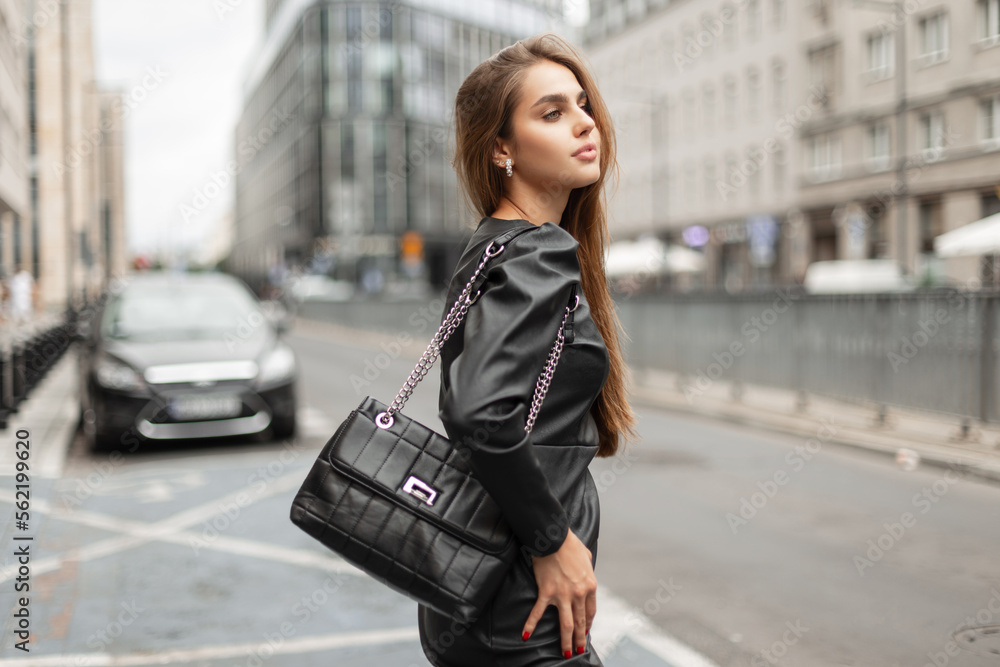 Stylish elegant beautiful business girl model in fashion black clothes with a leather black dress and a fashionable bag walks in the city Stylish elegant beautiful business girl model in fashion black clothes with a leather black dress and a fashionable bag walks in the city