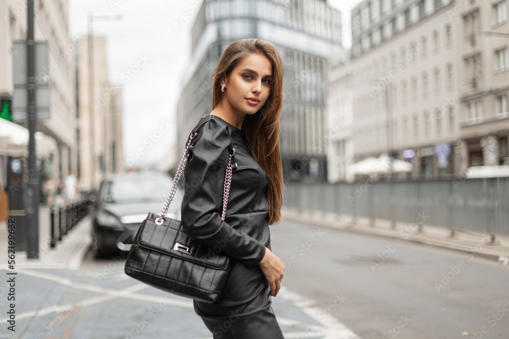Beautiful stylish business lady in fashionable black clothes with a dress and a fashion black leather bag is walking in the city. Urban elegant girl