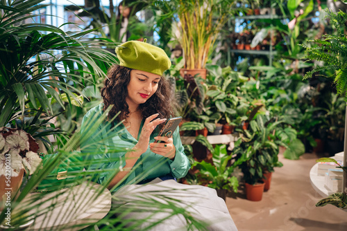 Stylish hipster woman using mobile phone in a cage with many green house plants. Hygge interior design. Biophilic lifestyle