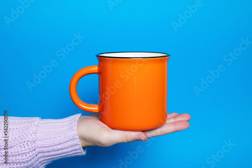A woman's hand holds a large orange cup with a hot drink on a blue background. The hand of a woman dressed in a pink sweater holds a ceramic cup on a bright blue background