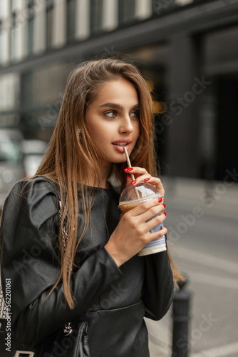 Beautiful stylish girl in fashion black clothes with a black leather dress and a bag walks in a modern city and drinking coffee