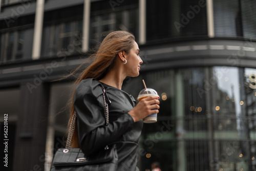 Successful beautiful stylish business woman in fashionable black clothes with a fashion leather bag walks in the city and drinks coffee against the background of a black glasses modern building