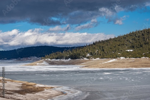 Winter panoramic landscape of mountain lake with frozen water and snow on the ground. Grey clouds on blue sky, coniferous forest. Natural widescreen view from Bulgaria, Rila mountains, Belmeken dam.