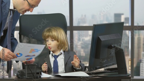 Photography Man bring paper work to little blond little boss, child in suit signing contract