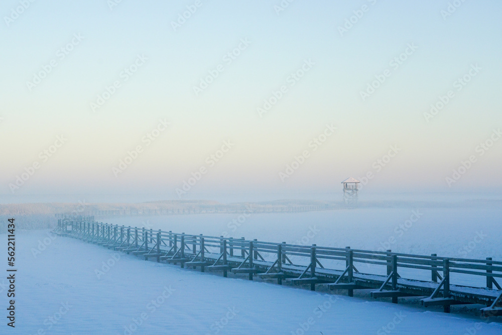 Wooden boardwalk on the frozen lake leading to the bird watching tower, winter sunset landscape