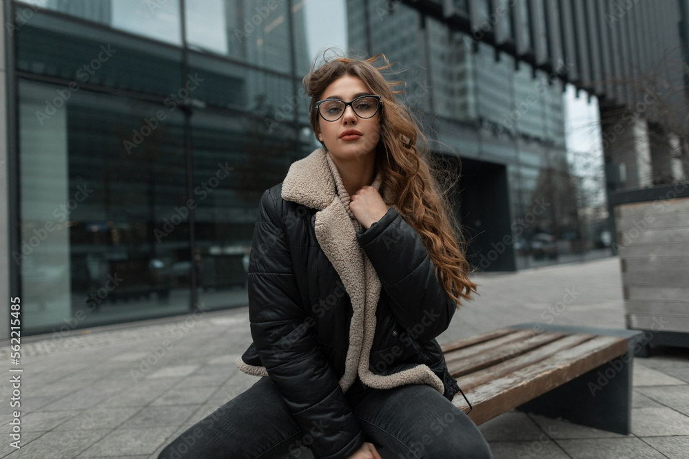 Fashionable beautiful young girl model with glasses in stylish winter clothes with a knitted sweater and a down jacket sits on a bench in the city