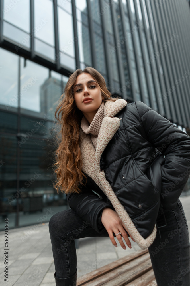 Beautiful fashionable woman model with a curly hairstyle in fashion winter clothes with a down jacket and a knitted sweater with jeans posing near a business building in the city Beautiful fashionable woman model with a curly hairstyle in fashion winter clothes with a down jacket and a knitted sweater with jeans posing near a business building in the city