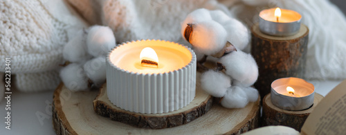 Cozy autumn or winter composition with aromatic candle, wool sweater, cotton, book. Aromatherapy, home atmosphere of cosiness and relax. Wooden background close up. Banner