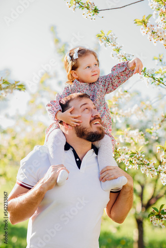 Dad plays and rolls on the shoulders his little daughter in a spring garden.