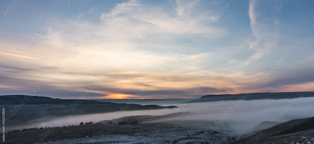 Obraz premium Winter panorama at sunset with snow covered hills with low fog in the valley and cloud cover, hills with snow and trees in winter at sunset