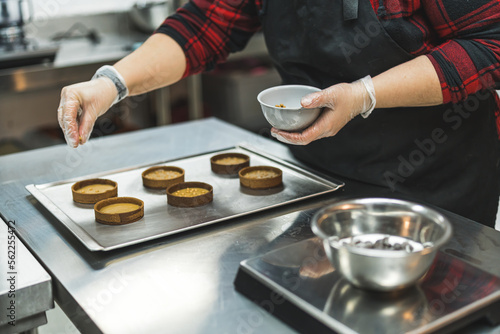 Female professional baker wearing black apron sprinkling golden sprinkles onto filled tart crusts. Baking process. Assembling dessert. Horizontal indoor shot. High quality photo