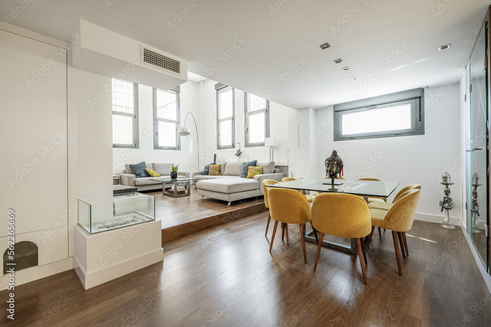 Double height loft apartment with square dining table with yellow fabric upholstered chairs and perimeter armchairs below windows