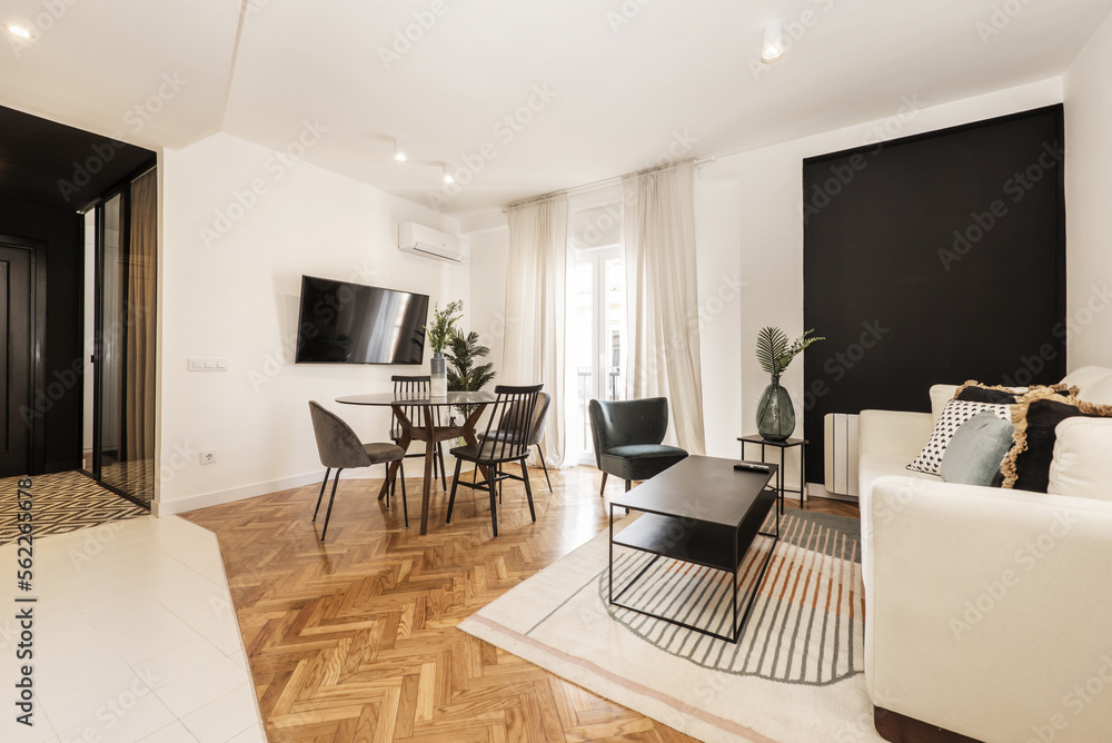 Fototapeta premium Loft room with round black glass dining table with vase and open plan kitchen in contemporary design, wall mounted tv, fabric sofa and black metal coffee table and balcony with sheers