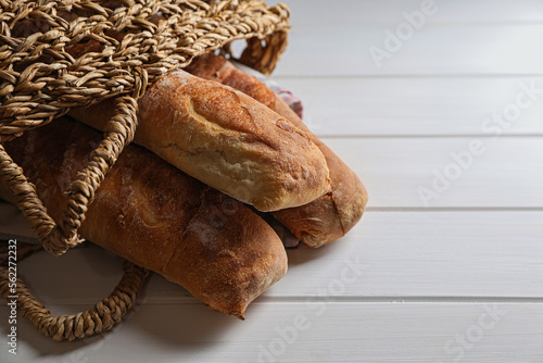Wicker bag with fresh crispy ciabattas on white wooden table, closeup. Space for text
