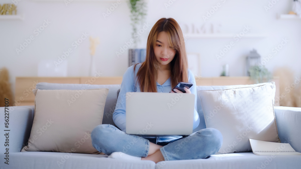 Young asian woman using computer laptop and mobile smartphone while seated on couch at home Young asian woman using computer laptop and mobile smartphone while seated on couch at home