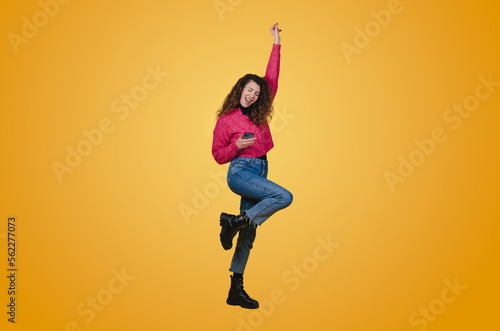 Portrait of a happy young woman celebrating victory holding one fist up while holding mobile phone isolated over yellow background. Happy people positive