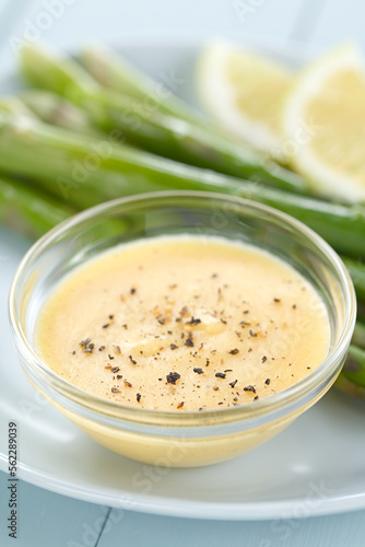 Hollandaise sauce with freshly ground black pepper served in glass bowl, green asparagus and lemon slices in the back (Selective Focus, Focus one third into the image)