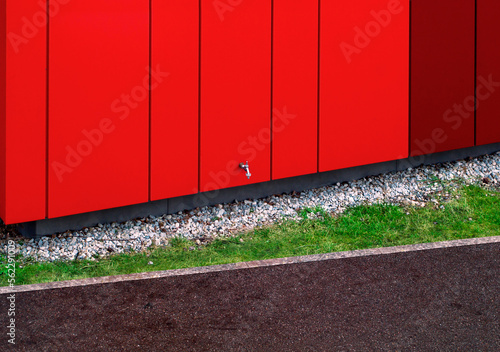 Water faucet in the wall of the house. Exterior view of the building facade with metallic red wall panels. White pebbles as drainage, green lawn and paver path with a curb.