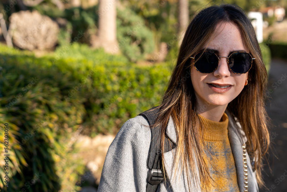 Young female in stylish outfit and sunglasses with backpack standing against blurred background of green bushes in sunny weather Young female in stylish outfit and sunglasses with backpack standing against blurred background of green bushes in sunny weather