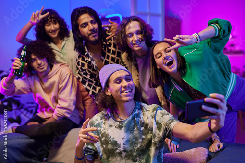 Group of young cheerful intercultural friends in casualwear taking selfie at home party while standing in living room lit by neon light